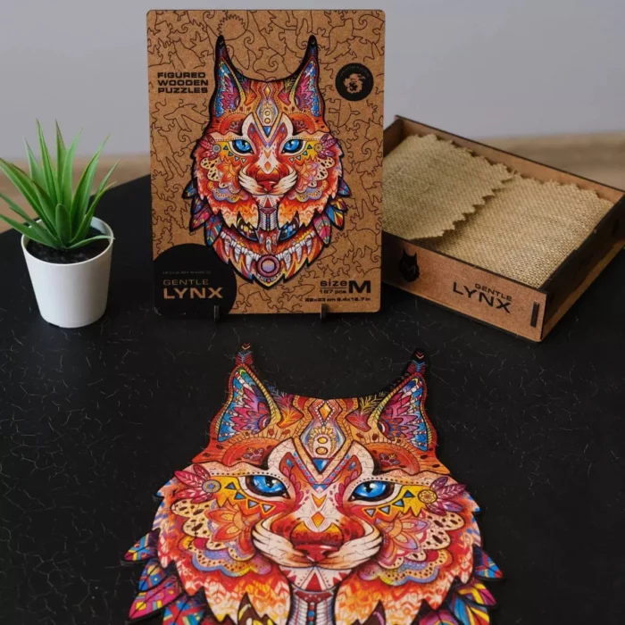 Un puzzle colorato raffigurante il volto stilizzato di Gentle Lynx Unidragon Animali disposto su un tavolo accanto alla sua scatola con una piccola pianta in vaso a lato.