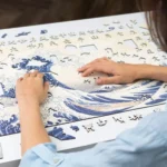 Donna che assembla un puzzle Unidragon Art LA GRANDE ONDA AL LARGO DI KANAGAWA raffigurante LA GRANDE ONDA di KANAGAWA su un tavolo.
