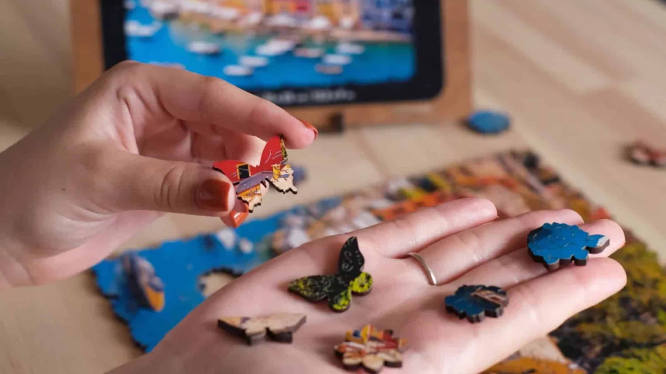 Mani che tengono i pezzi del puzzle sopra un tablet che mostra l'immagine del puzzle completato, con più pezzi sparsi su un tavolo di legno.