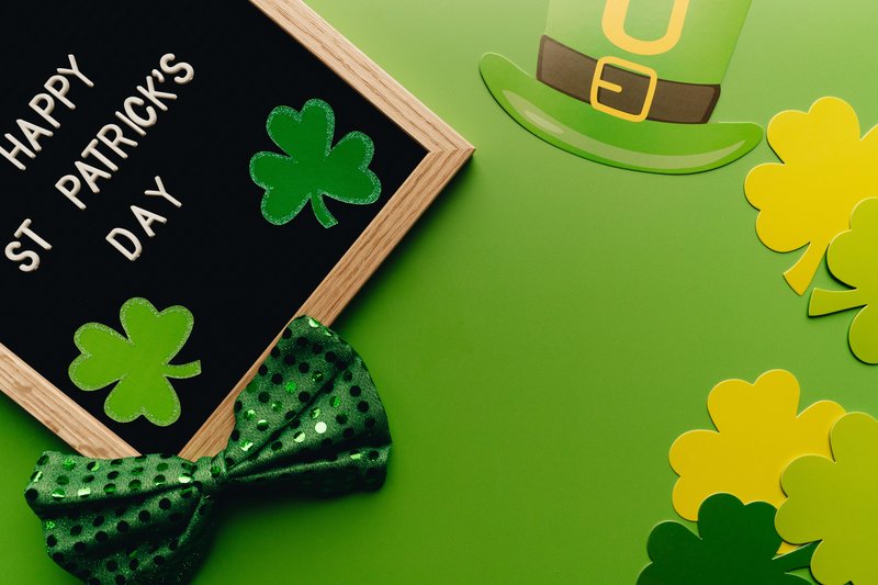 Composizione “flat lay” per il giorno di San Patrizio con una lavagnetta con lettere, trifogli scintillanti, un papillon verde, un cappellino di carta e sagome di trifogli su uno sfondo verde.