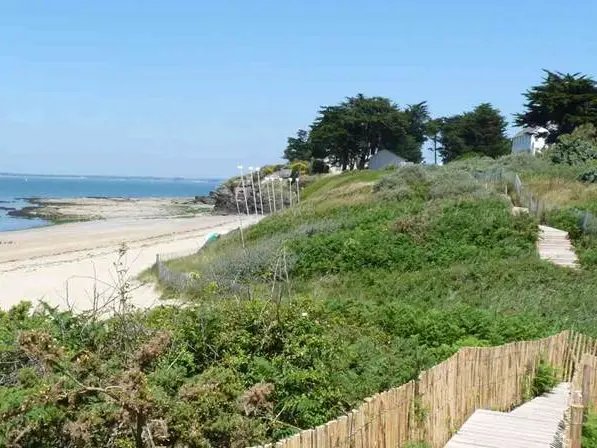 Una spiaggia sabbiosa con una passerella di legno, una vegetazione rigogliosa e alcune case su una collina erbosa affacciata sull’oceano, sotto un cielo azzurro e limpido.