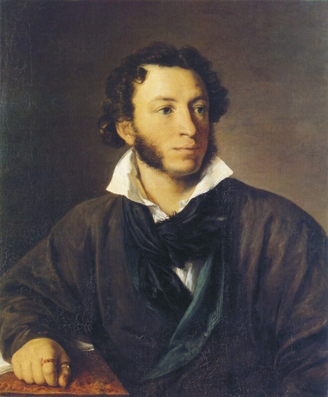 Un uomo con i capelli ricci, le basette e un cappotto scuro è seduto a un tavolo e guarda leggermente alla sua sinistra, su uno sfondo marrone a tinta unita.