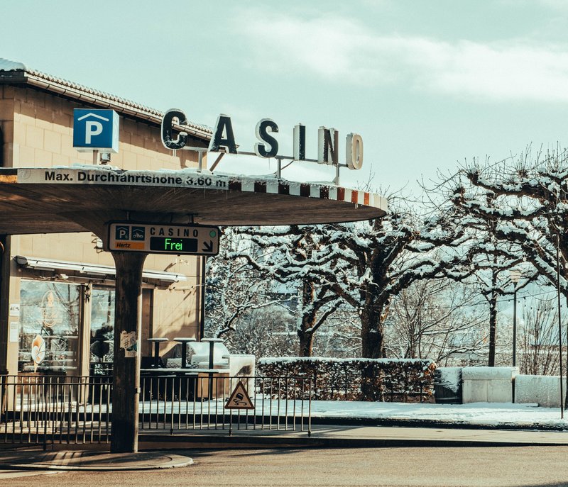 Ingresso di un casinò con un cartello che indica il parcheggio, alberi invernali e neve sul terreno. Un cartello recita “Frei”, a indicare che ci sono posti auto disponibili.