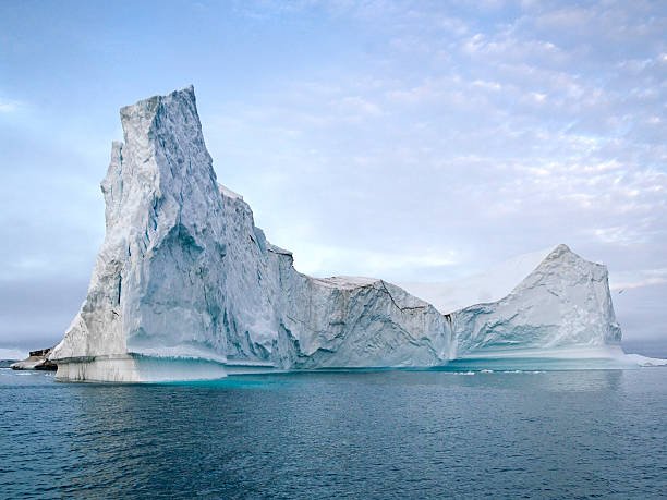 Un grande iceberg dalle cime frastagliate galleggia nelle calme acque blu dell'oceano sotto un cielo parzialmente nuvoloso.