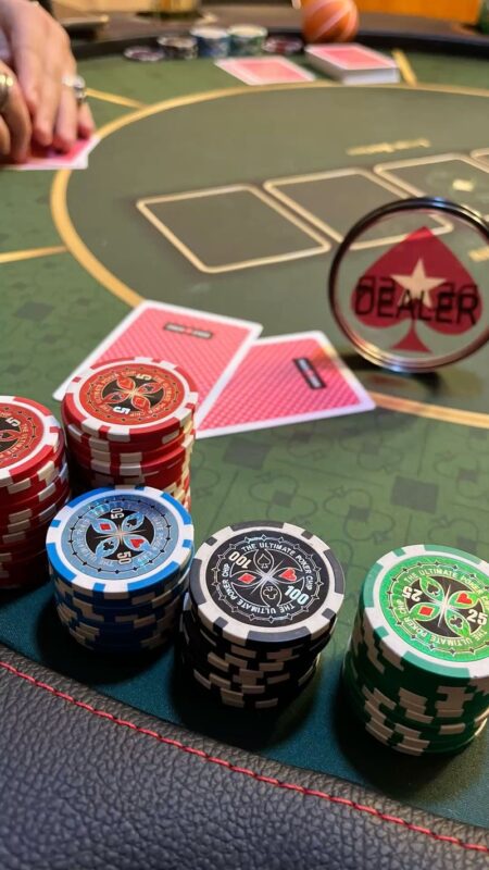 Su un tavolo da poker sono impilate fiches rosse, blu, nere e verdi. Le carte vengono distribuite a faccia in giù ed è visibile il bottone del “dealer”. Sullo sfondo, una persona tiene in mano delle carte.