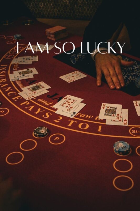 Un tavolo da blackjack con carte, fiches da poker e la mano di una persona in primo piano, con la scritta “I AM SO LUCKY.” sovrapposta.