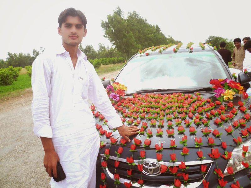 Un uomo in abito tradizionale bianco è in piedi accanto a un'auto Toyota nera decorata con fiori artificiali rossi e gialli, all'aperto in una giornata nuvolosa.