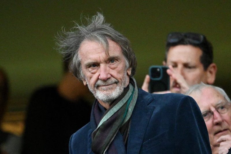 Un uomo anziano, con i capelli grigi arruffati e una sciarpa, è in piedi davanti a un gruppo di persone, una delle quali sta scattando una foto con uno smartphone.