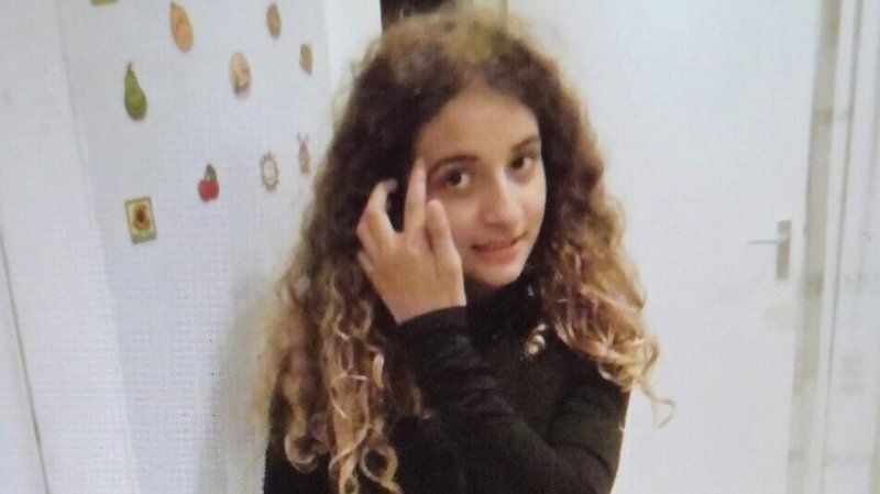 Una ragazzina con lunghi capelli ricci e una maglietta nera è in piedi in una stanza, mentre si tocca la fronte; alle sue spalle, su una parete bianca, si vedono degli adesivi a forma di frutta.