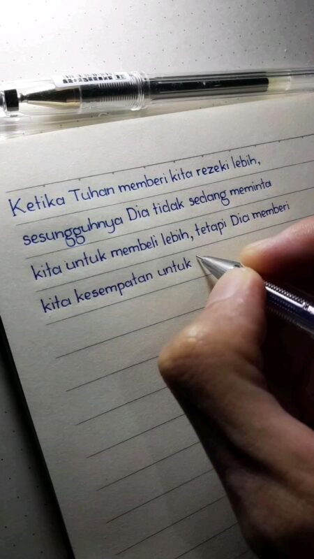 Una mano scrive una citazione motivazionale in indonesiano con una penna su un foglio a righe; una penna nera è appoggiata in cima alla pagina.