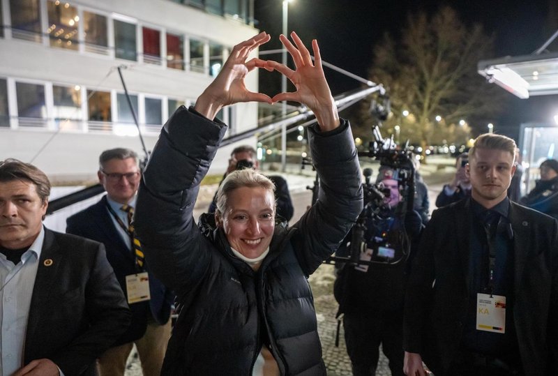 Una donna con un piumino forma un cuore con le mani mentre si trova all’aperto di notte, circondata da diverse persone e da attrezzature fotografiche.