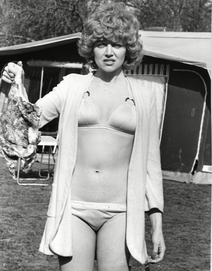 Una donna in bikini e vestaglia aperta è in piedi all'aperto e tiene in mano un pezzo di stoffa o un indumento con un'espressione scontenta. Sullo sfondo si intravede una tenda o una roulotte.