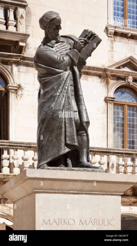 Una statua in bronzo raffigurante un uomo con un libro in mano è collocata su un piedistallo su cui è inciso il nome “Marko Marulić”, con sullo sfondo un edificio in pietra con finestre ad arco.